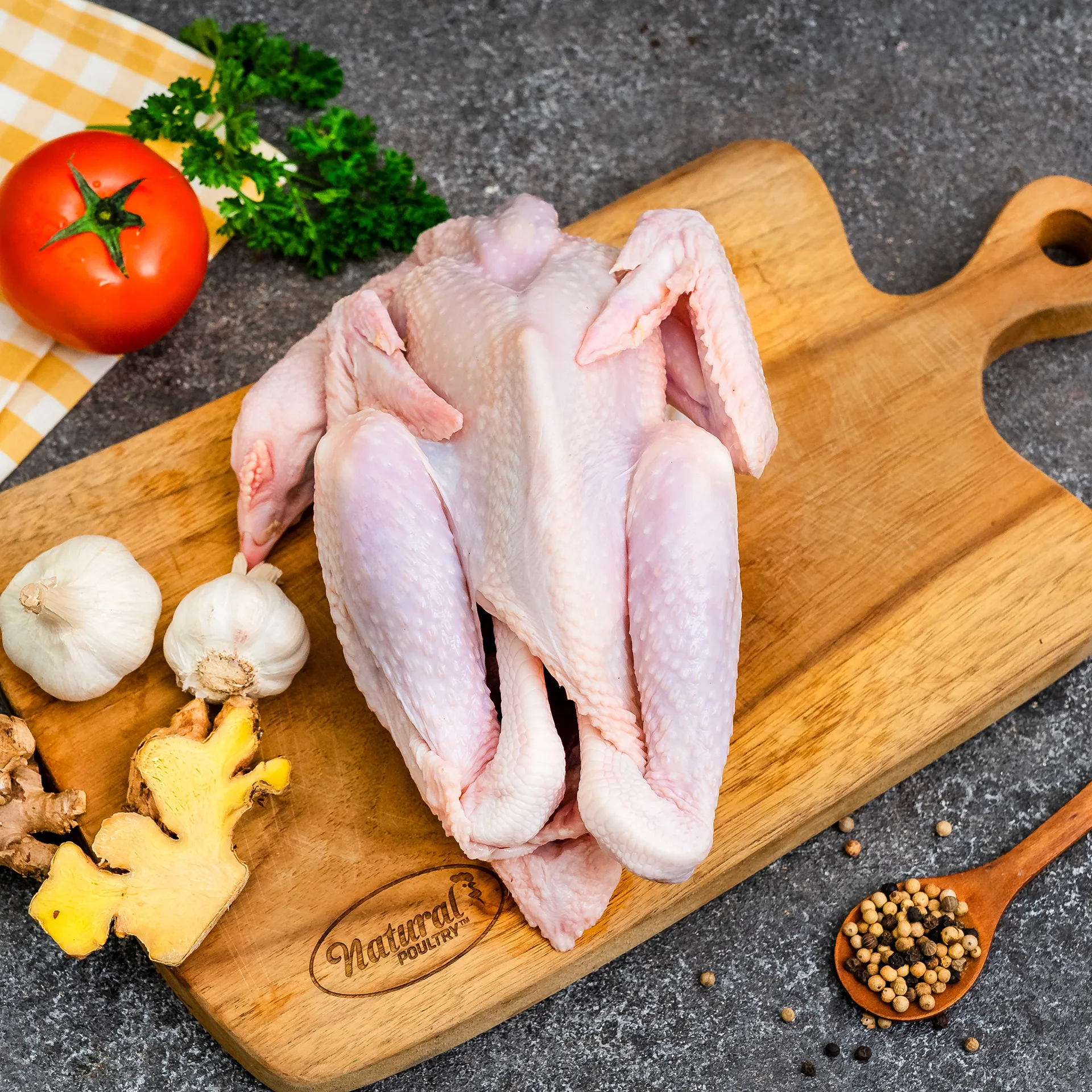 Foto produk ayam utuh di atas talenan kayu dengan bawang putih, jahe, dan tomat